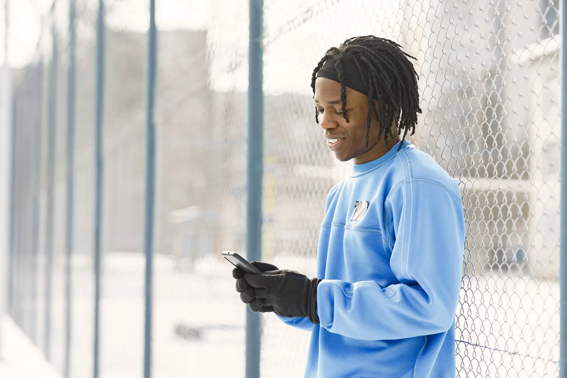 Smiling man with dreadlocks in blue sweater using smartphone by fence on sunny day.