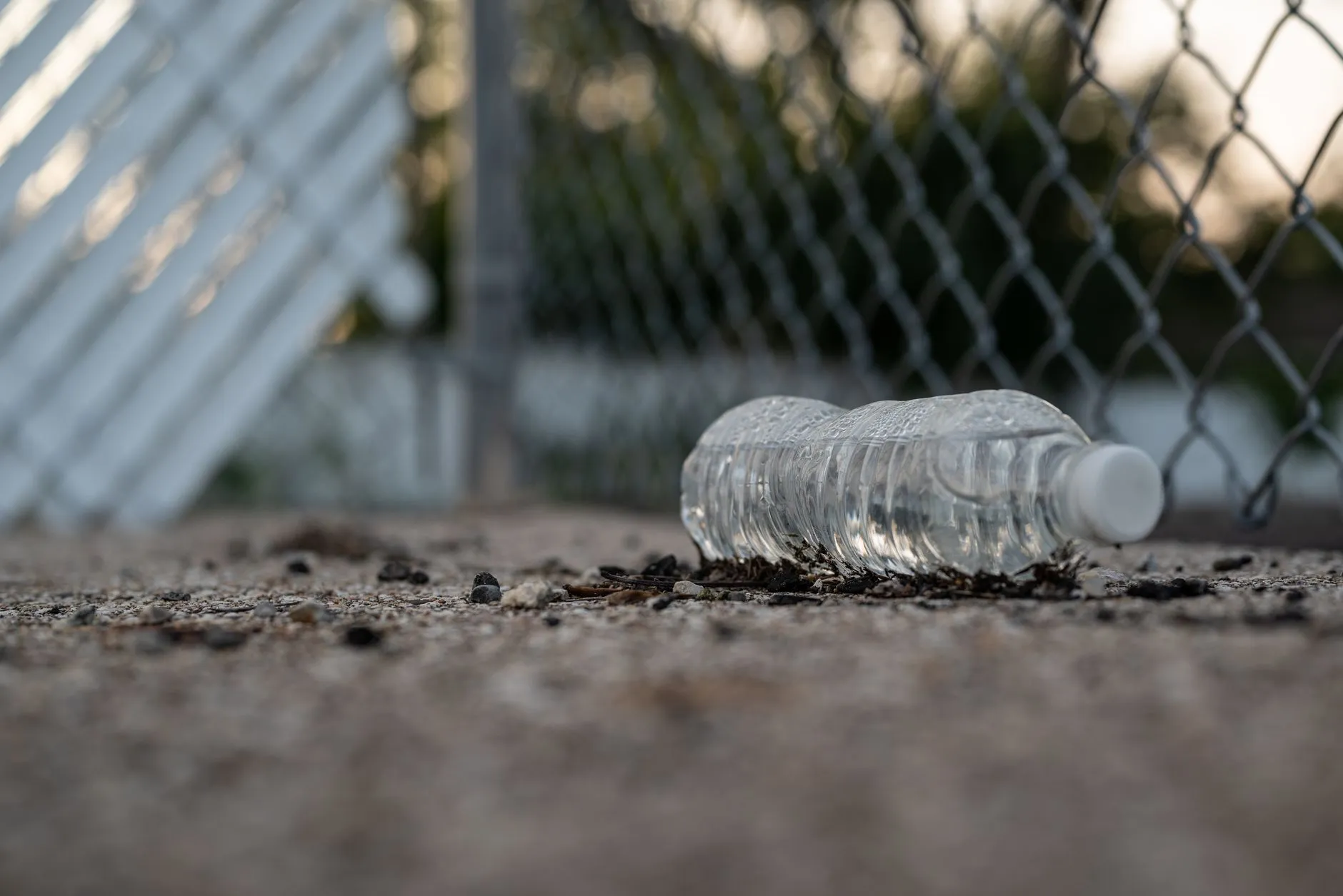 Close-up of a plastic water bottle discarded on gravel near a chain-link fence.